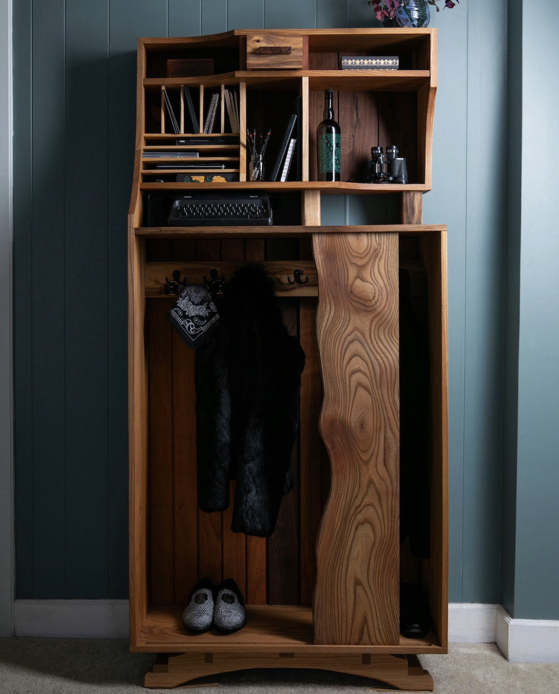 Entryway cabinet featuring a coat rack in its base and shelving in the upper portion of the piece. Personal effects are placed in furniture, such as shoes, a coat, stationary, and a typewriter.