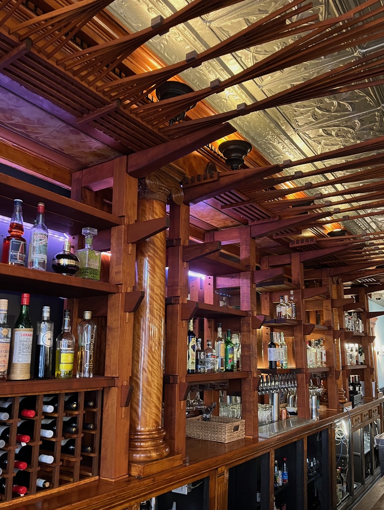 Bar shelving. Featuring front of shelving unit, with keyed mortises and tenons, and a wine rack. A walnut  array of thin slats artistically arrange on top of shelving is highlighted by overhead lighting. 