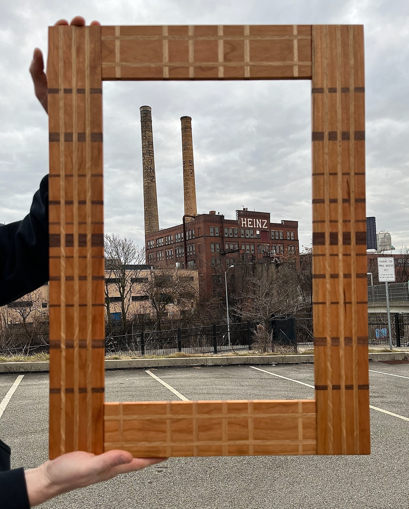 Cherry frame with white oak, ash and walnut inlays in a geometric linear pattern. Frame is being held by two hands in the foreground with the historic Heinz factory in the background. Troy Hill, North Side, Pittsburgh Pennsylvania. 