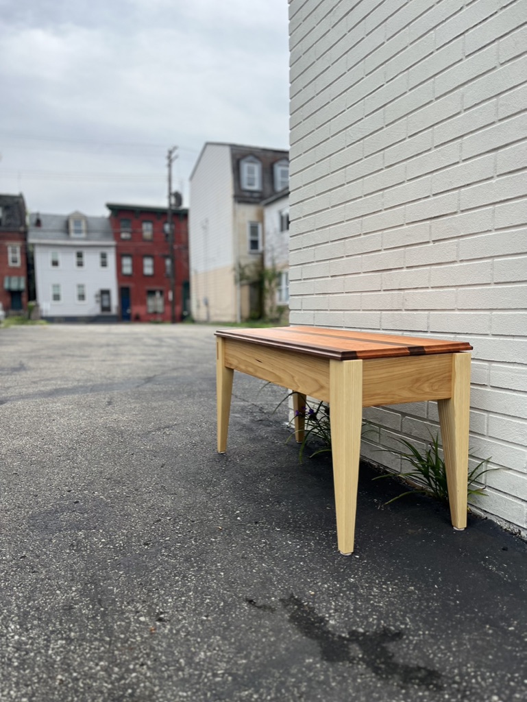 Piano bench, with lid closed, against a white brick wall in a neighborhood setting. Walnut, elm, ash, and cherry wood species.