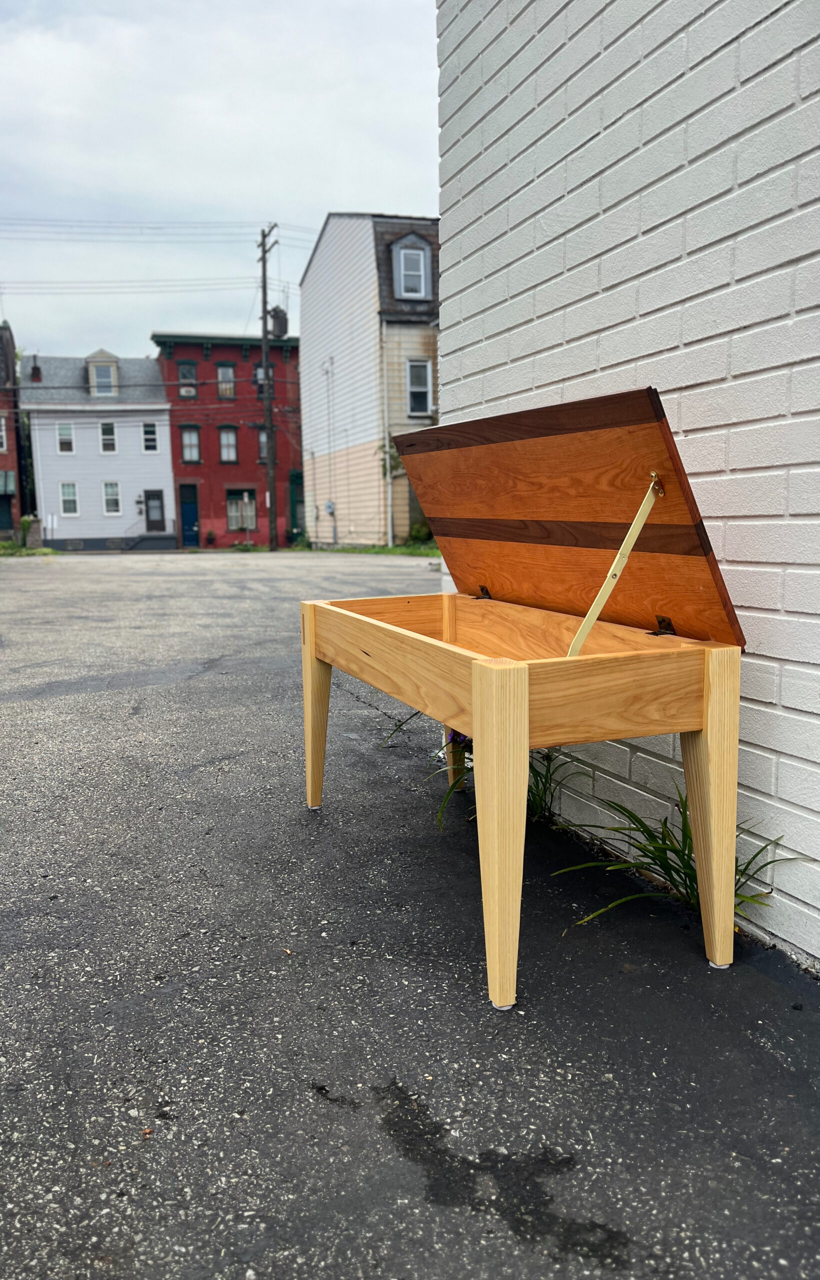 Piano bench, with lid open, against a white brick wall in a neighborhood setting. Walnut, elm, ash, and cherry wood species.