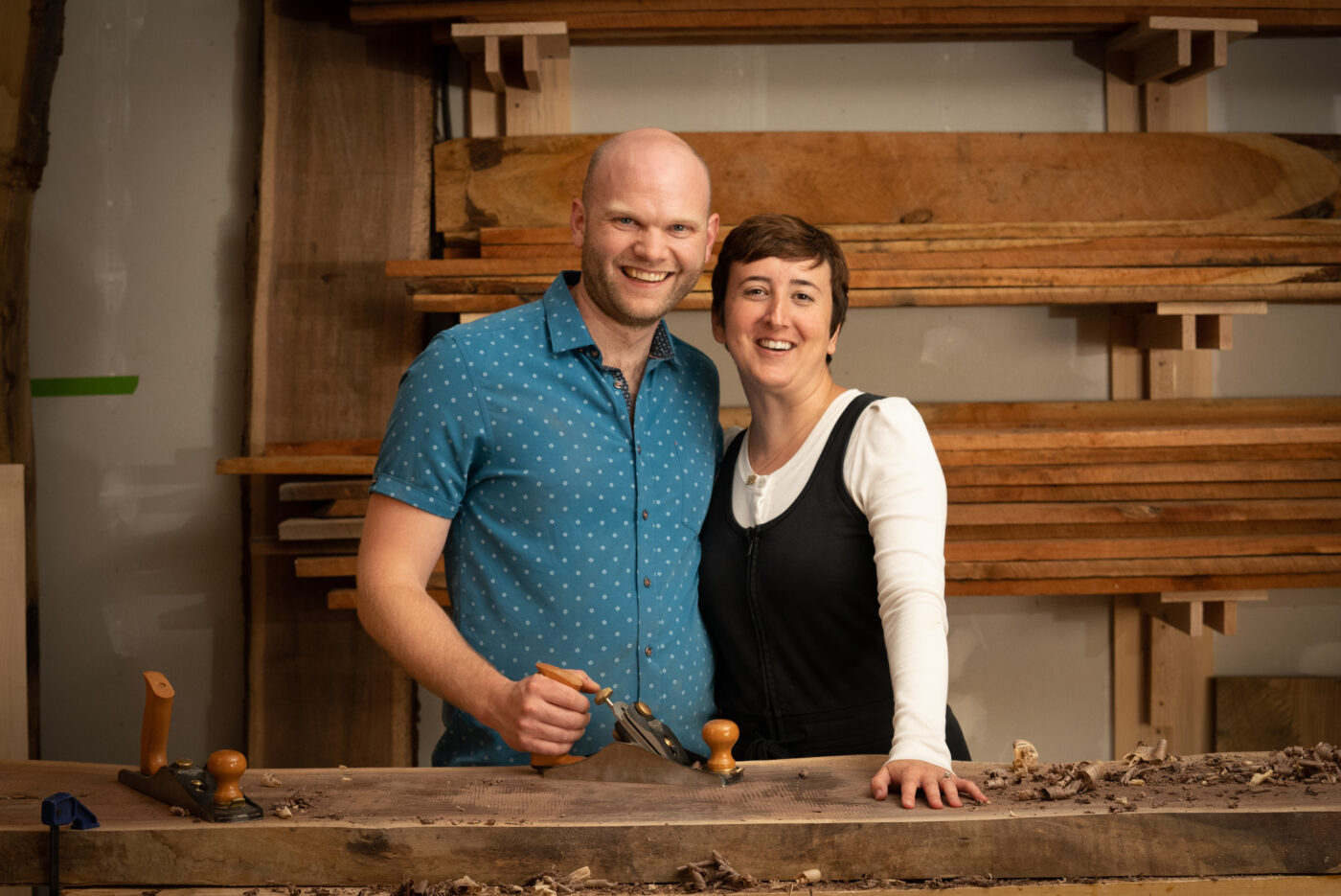 Lisa and Caleb Sisco smiling in their studio space. Caleb holds a wood planer with one hand that sits atop a walnut slab. Lisa places her hand on the wood.