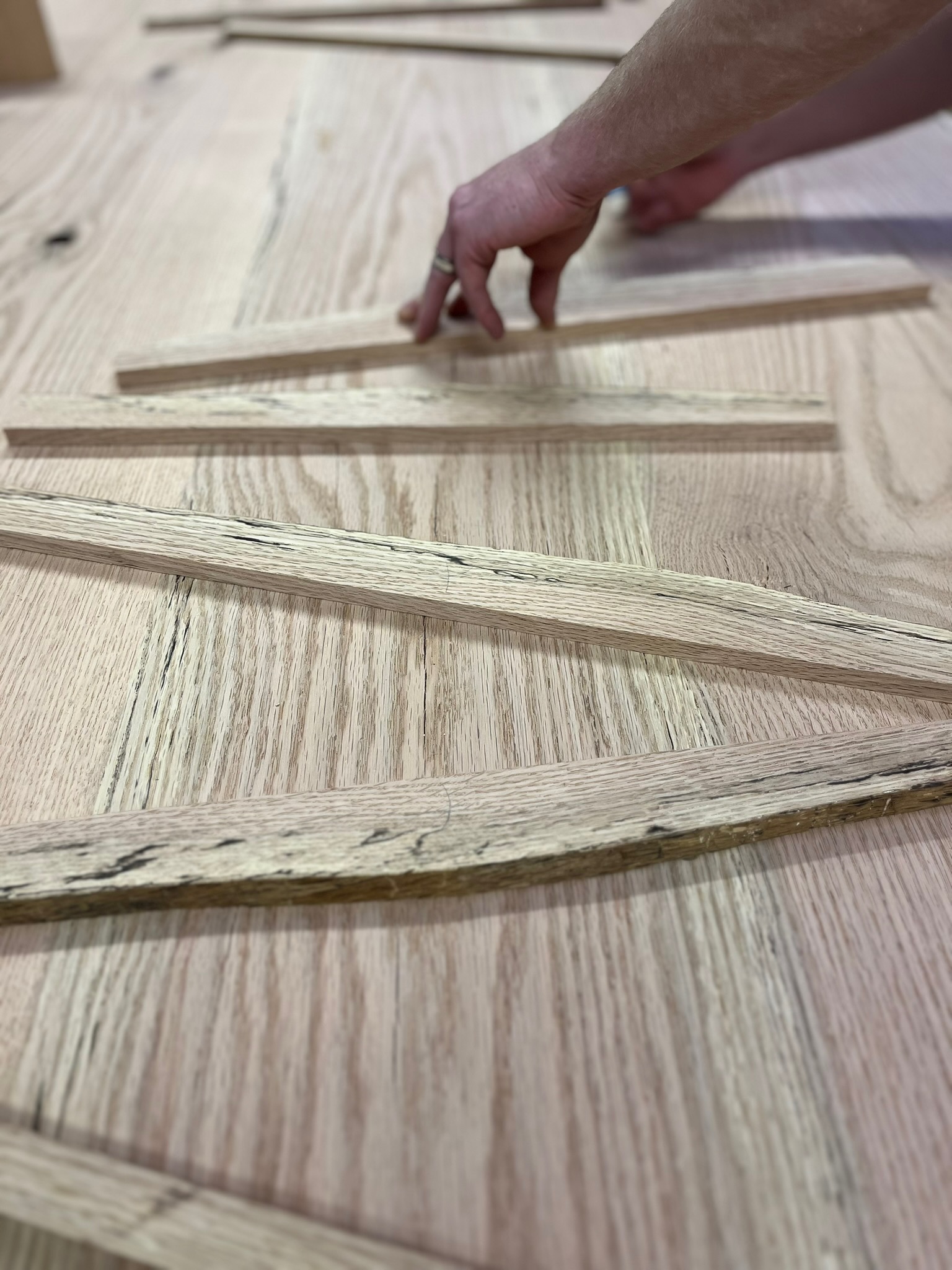 two hands and alternating thin strips of red oak wood on an in-progress tabletop. the strips of wood are spalted red oak and the table below is a pink hue, which is naturally occuring in red oak. 